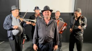 group of men in black shirts and hats playing violins, accordians and smiling at camera 